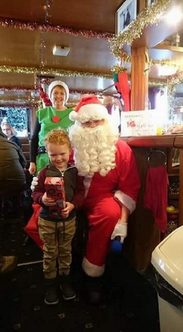 child with present standing next to santa with elf in background on board festive cruise boat