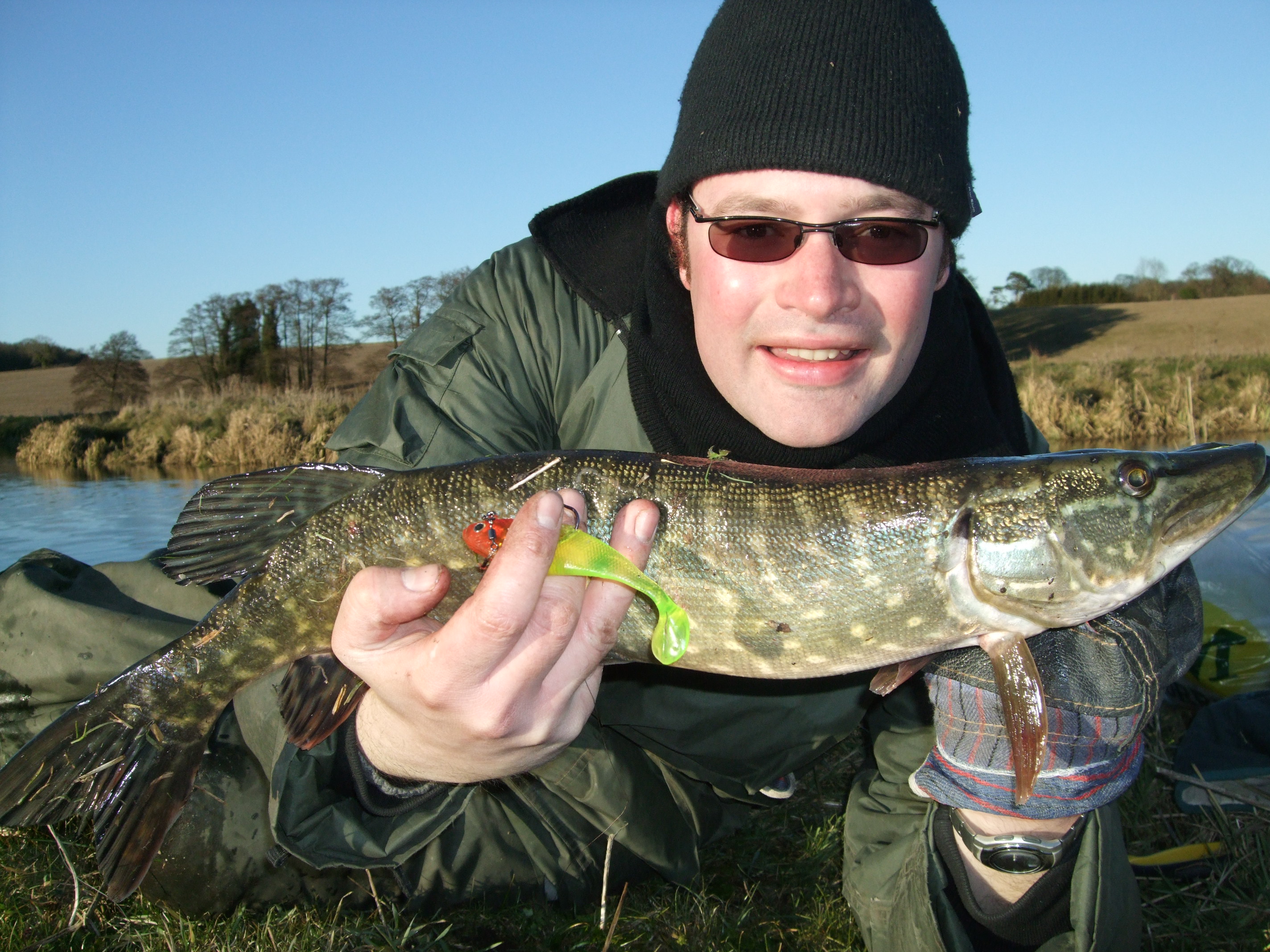 man wearing hat and sunglasses in a boat is holding a pike up to the camera